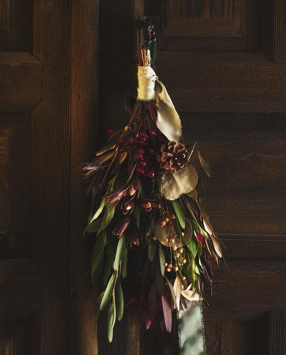 Dekorativna božična veja z zelenimi oljčnimi listi, storži in ratanovo pentljo, obešena na lesenih vratih.