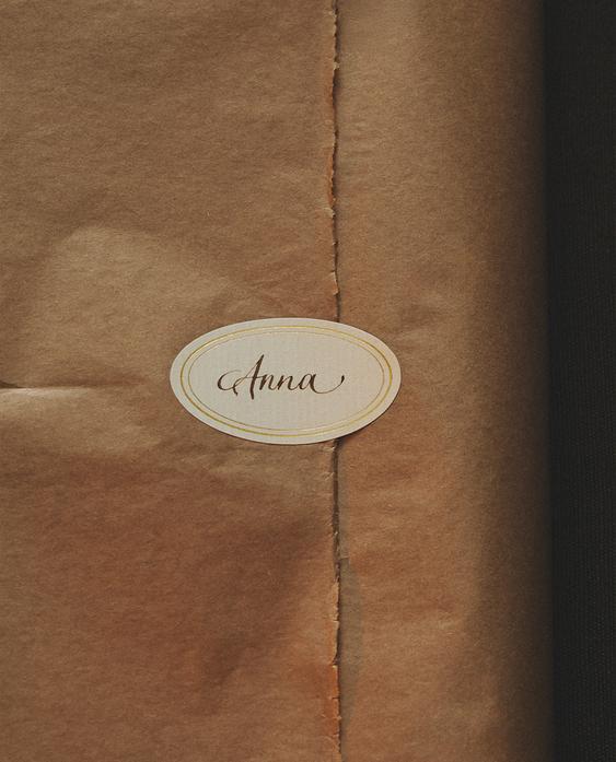 Podrobnost ovalne samolepilne etikete z napisom "Anna" na rjavem papirju.