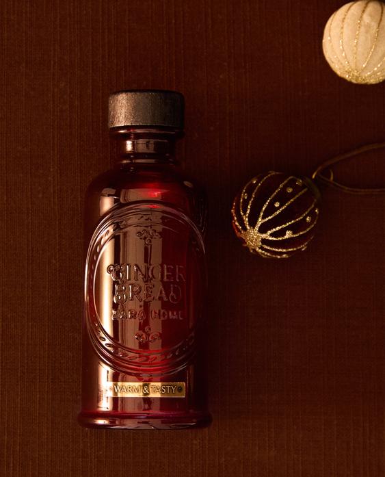 Rdeča steklenička difuzorja z napisom "GINGER BREAD ZARA HOME" in "WARM & TASTY" v reliefu, skupaj z božičnimi okraski.