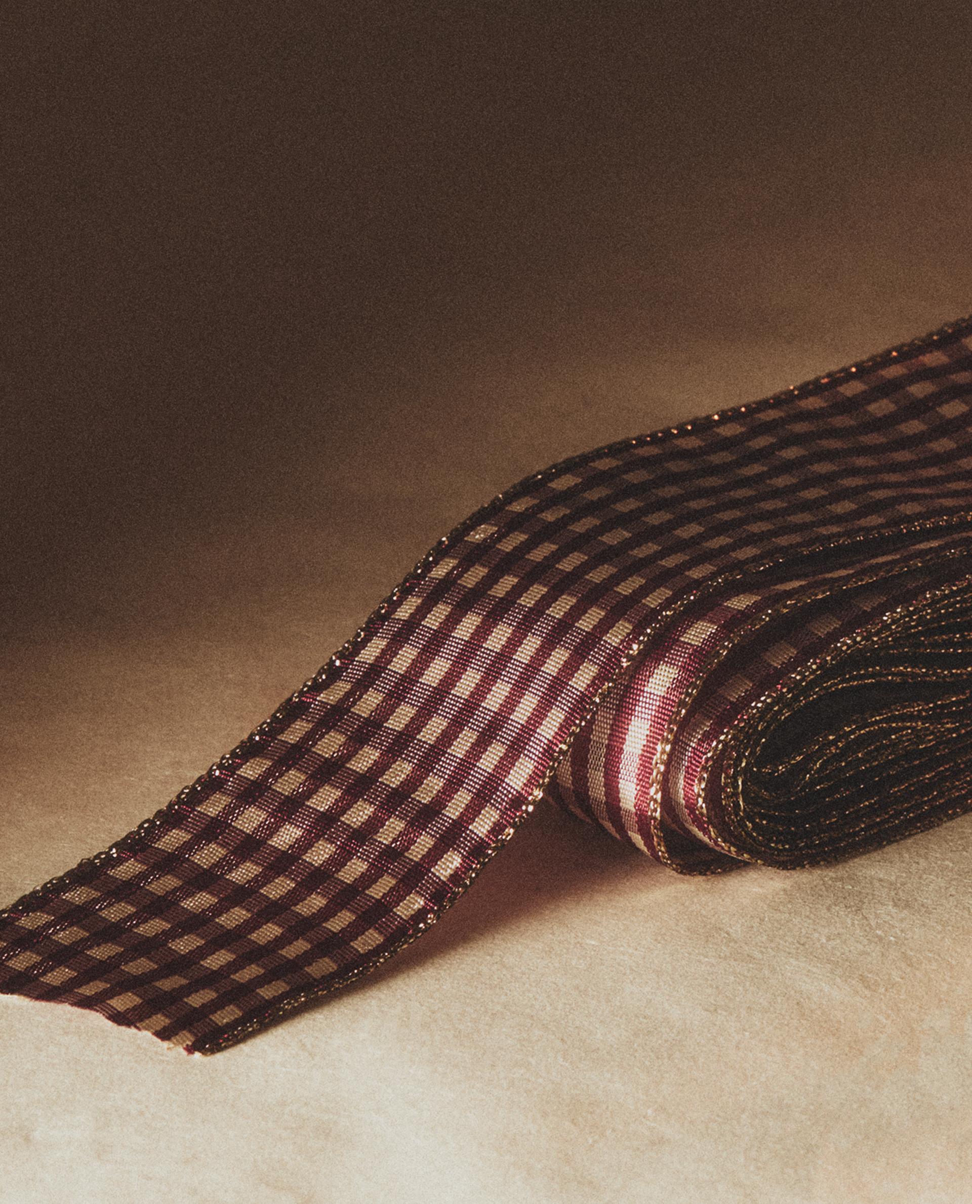 Rdeč trak z majhnim karirastim vzorcem in zlatimi robovi, delno zvit na bež površini.