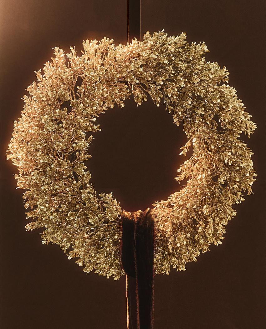 Okrasna zlata božična krona s sijočimi listi, obešena s temnim trakom. Približni premer 40 cm.