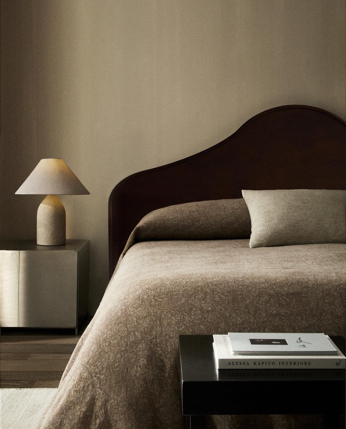 Colcha marrón con diseño jacquard floral sobre cama, acompañada de cojín a juego y lámpara en mesita lateral, MORRIS & CO. × ZARA HOME.