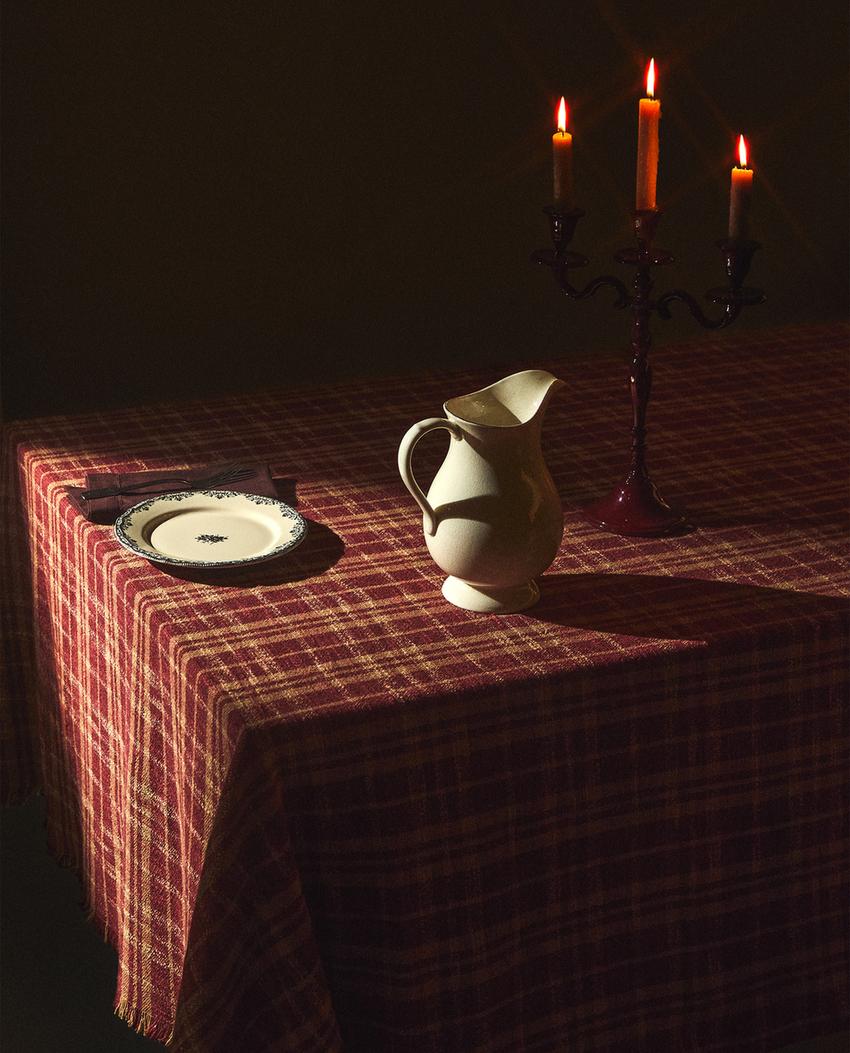 Sprednji pogled na karo prt v naravni in rdeči barvi na mizi, z belim vrčem, svečnikom in okrasnim krožnikom.