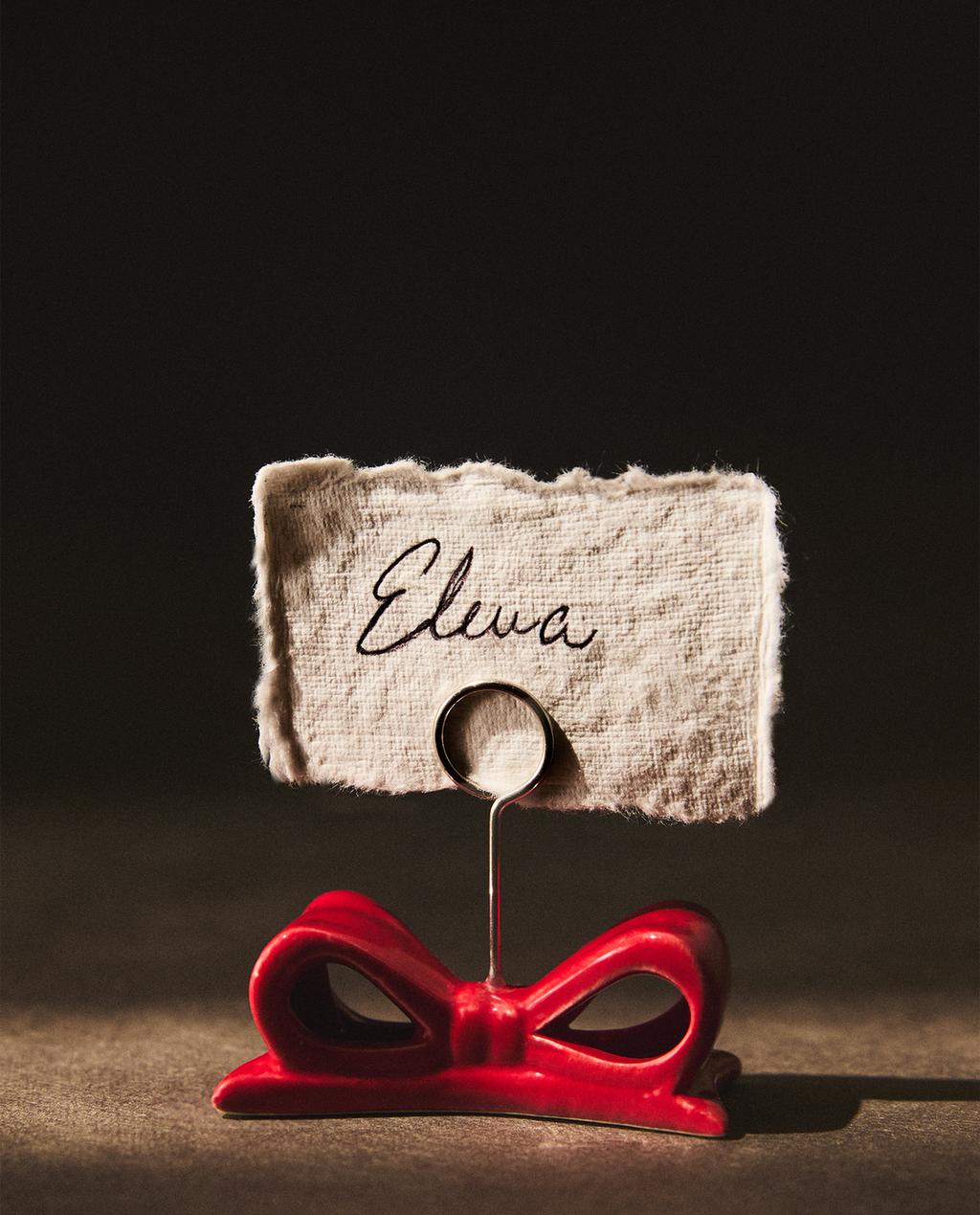 Rdeč markazit v obliki božične pentlje, zgornji obroč drži kartico z besedilom "Elewa".