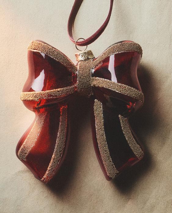 Božični okrasek v obliki rdeče pentlje z zlatimi bleščicami in trakom za obešanje.