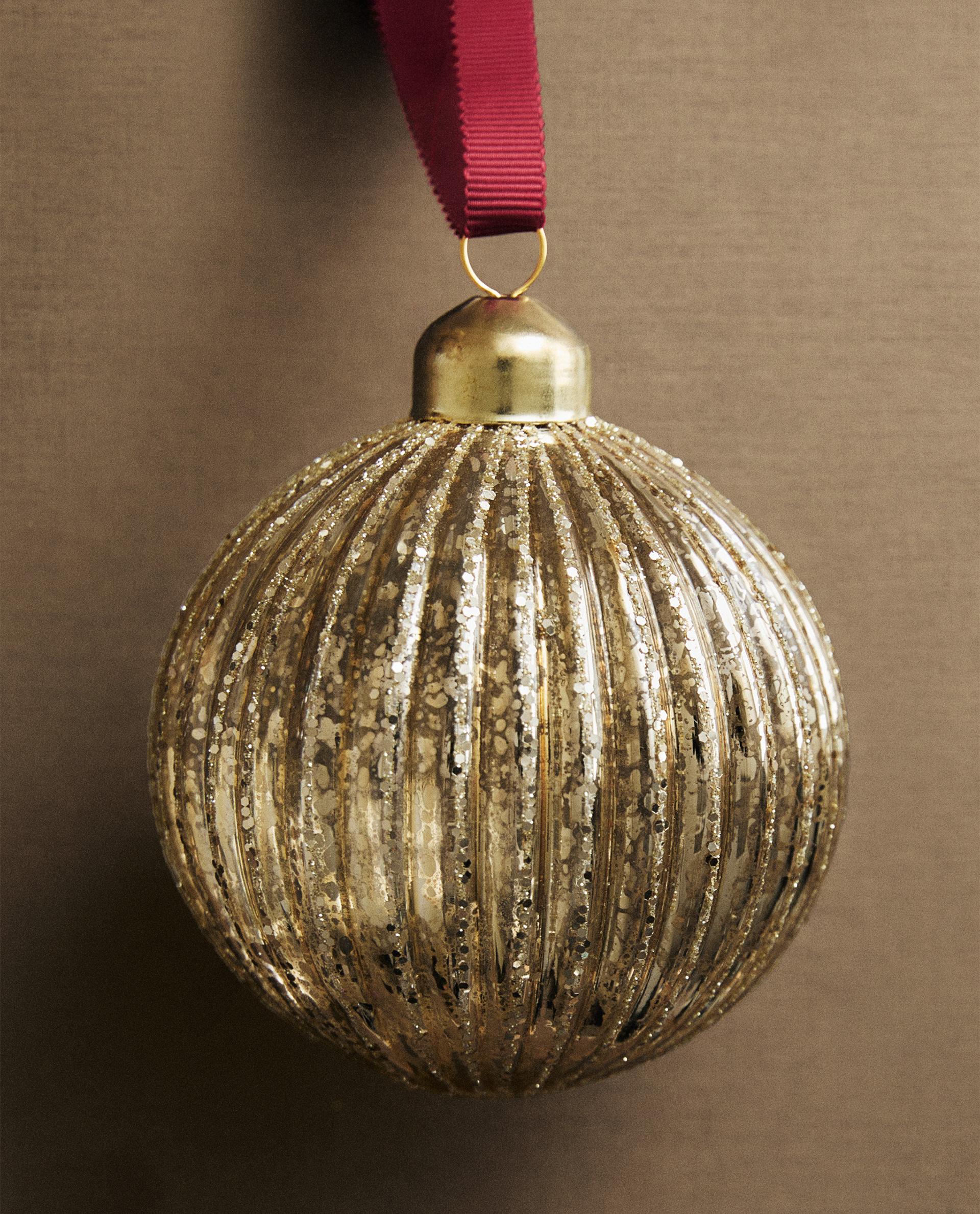 Zlati božični okrasek v obliki krogle s črtami bleščic, obešen z rdečim trakom.