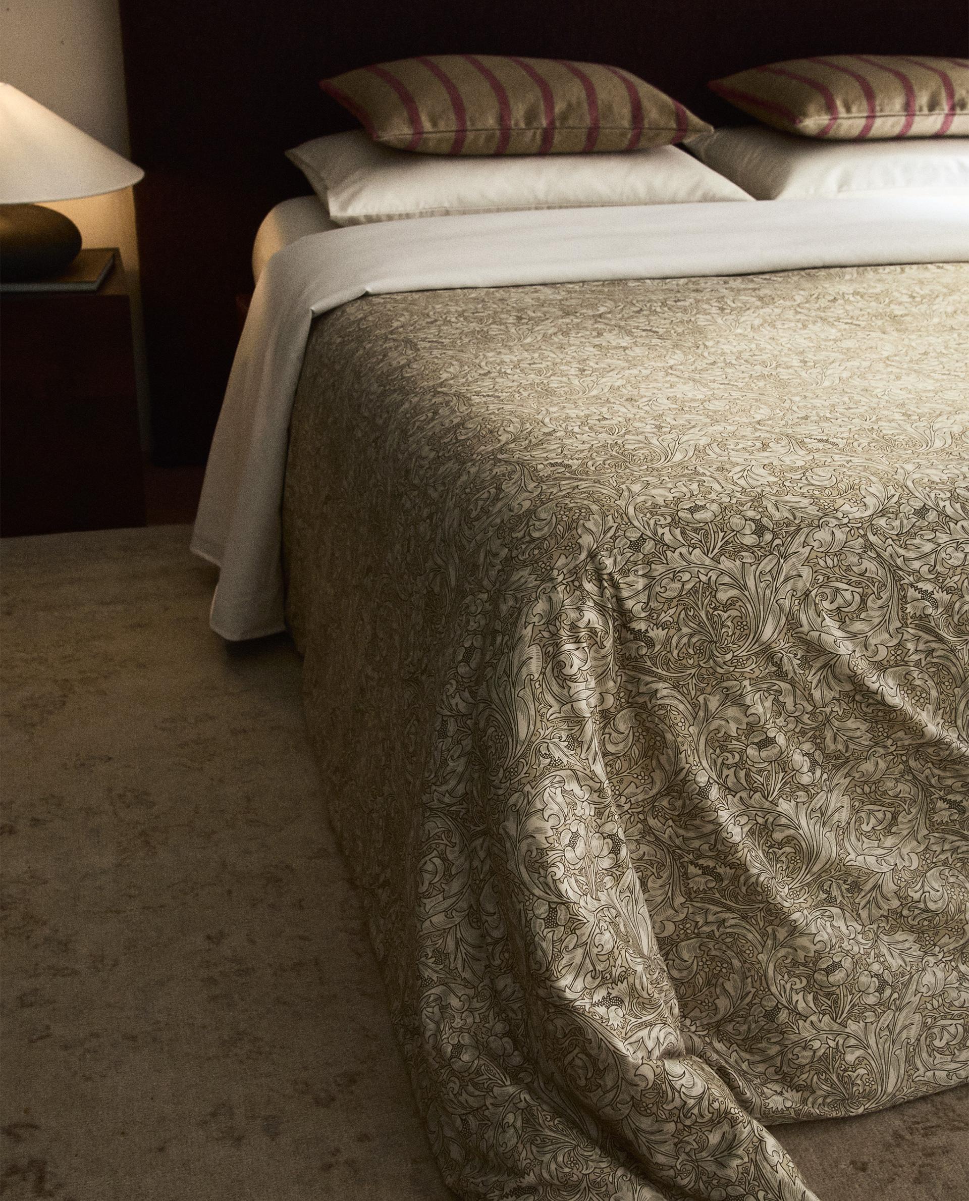Colcha con estampado floral en tonos beige y marrón sobre cama, acompañada de almohadas con fundas a rayas, MORRIS & CO. × ZARA HOME.