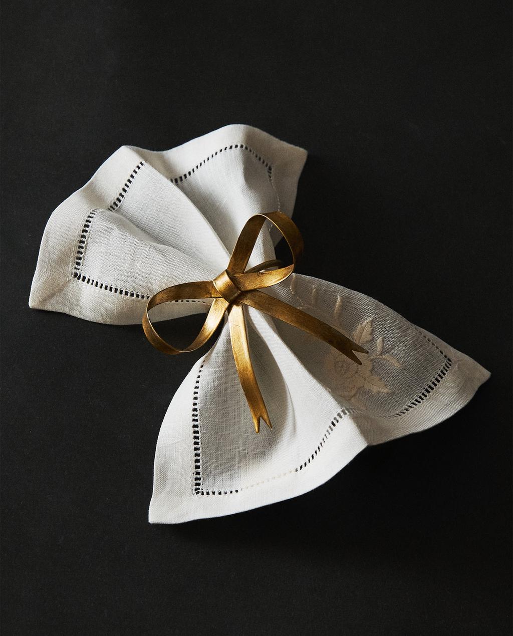 Zlat prstan za serviete z zasnovo pentlje, na beli servieti z okrasnimi robovi.